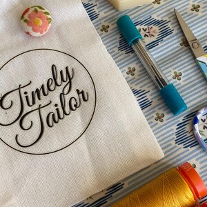May include: A white fabric bag with the words "Timely Tailor" printed in black. The bag is sitting on a blue and white patterned fabric with a spool of orange thread, a spool of yellow thread, a pair of scissors, a small container of wax, and a floral button.