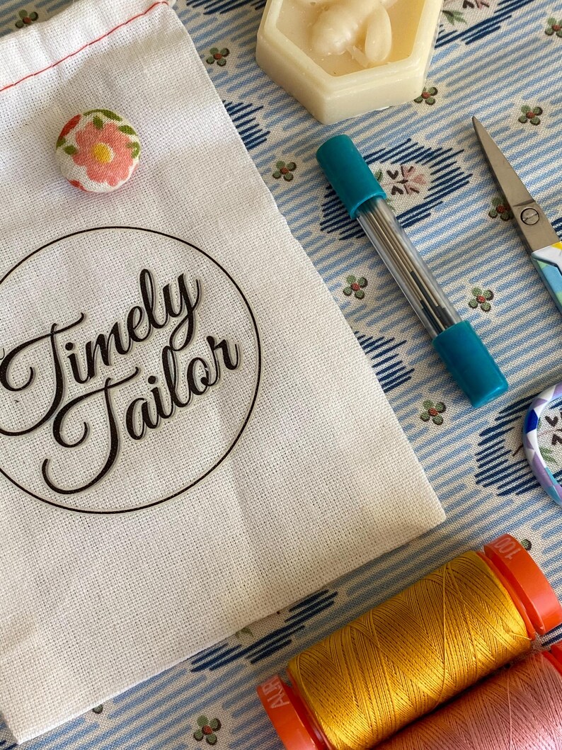 May include: A white fabric bag with the words "Timely Tailor" printed in black. The bag is sitting on a blue and white patterned fabric with a spool of orange thread, a spool of yellow thread, a pair of scissors, a small container of wax, and a floral button.