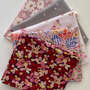 Peut inclure: Une pile de cinq carrés de tissu pliés aux motifs variés. La pièce supérieure est bordeaux foncé avec des feuilles roses et des fleurs jaunes. Les autres couches incluent un imprimé floral rose clair, un tissu gris avec des épingles blanches.