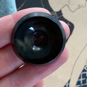 May include: A black, glossy button with two holes. The button has a slightly concave design and a reflective surface. The button is round and appears to be made of a hard material.