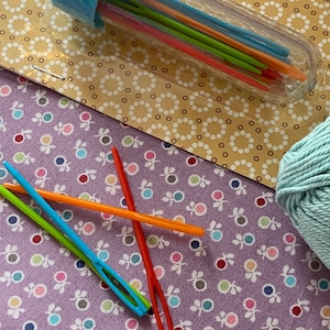 May include: A clear plastic container filled with colorful plastic needles. The container has a blue lid and is sitting on a purple fabric with a white polka dot pattern.