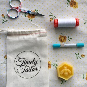 May include: A white cotton drawstring bag with the text "the Timely Tailor" printed in black. The bag is next to a spool of white thread, a pair of colorful scissors, a blue pen, and a yellow beeswax block with a bee design.