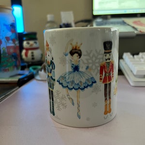 May include: White ceramic mug with a winter-themed design featuring a ballerina, a nutcracker, and snowflakes. The ballerina is wearing a blue and white dress and is holding a crown.