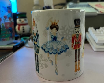 May include: White ceramic mug with a winter-themed design featuring a ballerina, a nutcracker, and snowflakes. The ballerina is wearing a blue and white dress and is holding a crown.