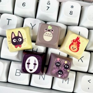 Op de afbeelding: Een wit toetsenbord met kleurrijke toetsen met daarop karakters uit Studio Ghibli-films. De toetsen bevatten No Face, Totoro, Jiji en personages uit Kiki's Bezorgdienst.