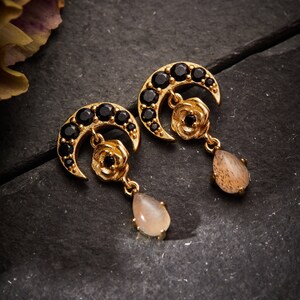 May include: A pair of gold earrings with a crescent moon design, black gemstones, and a flower charm. Each earring has a teardrop-shaped, light brown gemstone dangling from the bottom.
