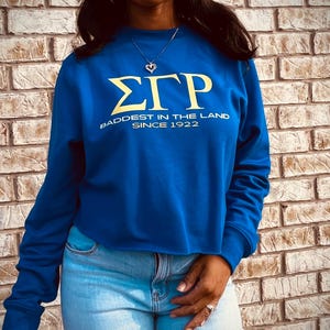 May include: A royal blue cropped sweatshirt with the letters "ΣΓΡ" in yellow and the text "Baddest in the Land Since 1922" in yellow.