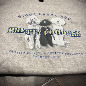 May include: A gray t-shirt with a blue and yellow graphic of three poodles. The text "Sigma Gamma Rho" and "Pretty Poodles" are printed on the shirt. The text "Greater Service - Greater Progress Founded 1922" is also printed on the shirt.