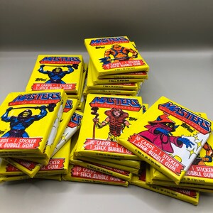 Pile of sealed 1984 Topps Masters of the Universe trading card wax packs with various character art including He-Man, Skeletor, and Orko. Vintage yellow Topps wrappers stacked neatly, emphasizing collectible condition.