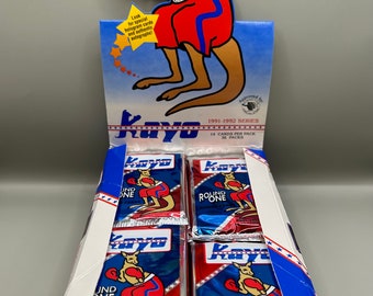 Puede incluir: Una caja de cartas de boxeo Round One de la serie 1991-1992 con un canguro de dibujos animados que lleva guantes de boxeo. La caja contiene 36 paquetes con 14 cartas por paquete.