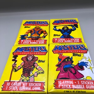 Front view of four sealed 1984 Topps Masters of the Universe wax packs featuring He-Man, Skeletor, Evil-Lyn, and Orko artwork on yellow wrappers. Each pack contains 10 cards, 1 sticker, and 1 stick of bubble gum.