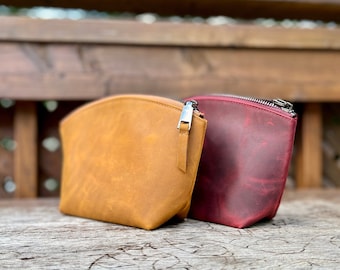 Könnte beinhalten: Zwei kleine Reißverschlusstaschen aus Leder, eine in senfgelb und die andere in einem tiefen burgunderrot. Die Taschen liegen auf einer Holzoberfläche.