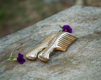 Könnte beinhalten: Zwei Holzkämme mit abgerundeten Griffen liegen auf einer verwitterten Holzoberfläche, akzentuiert durch kleine violette Blumen. Die Kämme haben feine Zähne und eine natürliche Holzmaserung, was auf natürliche Haarpflege hindeutet.