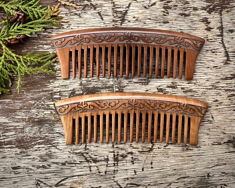 May include: Two wooden combs with intricate carved designs. The combs are brown and have a natural wood finish. The combs are laid out on a wooden surface.