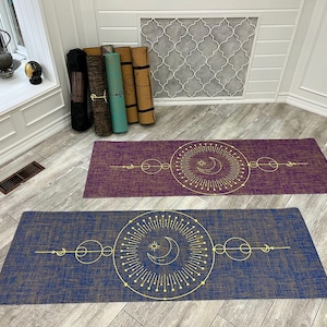 May include: Two yoga mats with a geometric design featuring a moon and sun symbol. The mats are made of natural jute and have a textured surface. The top mat is purple and the bottom mat is blue.