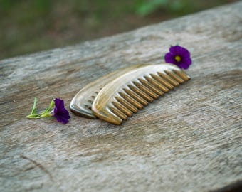 Könnte beinhalten: Zwei Holzkämme mit feinen Zähnen liegen auf einer verwitterten Holzoberfläche, begleitet von zwei lila Blumen. Die Kämme sind hellbraun und haben eine glatte Oberfläche, mit einer Länge von etwa 15 cm.
