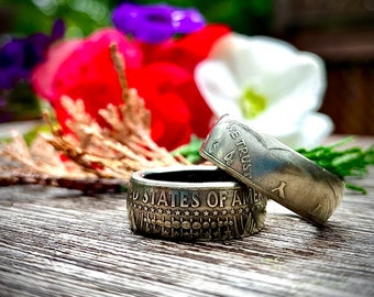 Könnte beinhalten: Zwei silberne Ringe, die aus US-amerikanischen 50-Cent-Münzen gefertigt sind. Der obere Ring trägt die sichtbaren Worte "In God We Trust" und "Liberty". Der untere Ring trägt die sichtbaren Worte "United States of America". Die Ringe liegen auf einer Holzoberfläche mit einem verschwommenen Hintergrund aus roten, weißen und blauen Blumen.