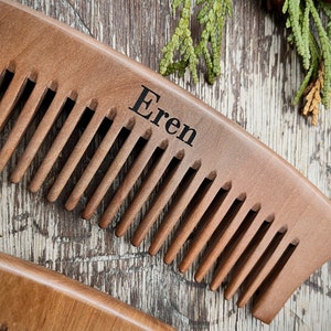 May include: A wooden comb with a curved handle and the name "Eren" engraved on the top. The comb is brown and has a natural wood finish.