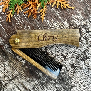 Peut inclure: Un peigne pliant en bois avec le nom "Chris" gravé sur le manche. Le peigne est ouvert, révélant des dents noires. Le bois a un grain naturel et est fixé avec une vis en laiton. Du feuillage vert et brun est en arrière-plan.