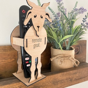 May include: A wooden remote control holder shaped like a goat. The goat is standing on a wooden shelf. The holder has the text "remote goat." on the front.