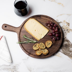 May include: A round wooden serving board with a handle, featuring a wedge of cheese, a bunch of grapes, rosemary sprigs, and toasted bread.