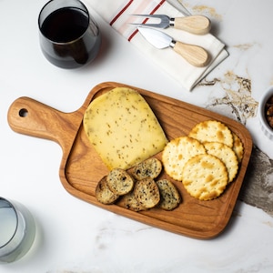 May include: A wooden cheese board with a handle holds a wedge of cheese, crackers, and toasted bread. Two glasses of wine and a small bowl of nuts are also present. Two cheese knives rest on a white cloth.