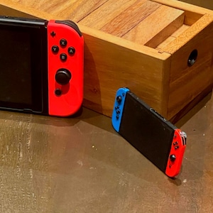 May include: A black handheld gaming console with blue and red controllers rests on a brown surface. A smaller version of the console is also present. A wooden box with circular cutouts is in the background.
