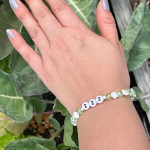 May include: A white beaded bracelet with green and white beads. The bracelet has the number 222 on it.