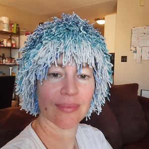 May include: A person wearing a blue and white fuzzy hat with long strands of yarn. The hat is covering the person's hair and forehead. The person is looking at the camera and smiling.