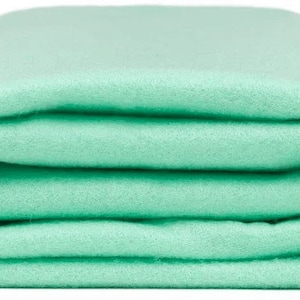 Puede incluir: Una manta de forro polar de color verde claro doblada. La manta es suave y mullida, perfecta para acurrucarse en el sofá.