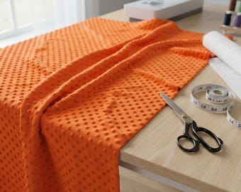 Puede incluir: Una tela minky de color naranja brillante con una textura de puntos en relieve drapeada sobre una superficie de madera clara. Junto a la tela hay unas tijeras de costura de metal con mangos negros y una cinta métrica blanca con marcas en cm.