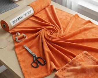 Puede incluir: Un rollo de tela minky de color naranja sólido con una etiqueta que dice SOLID MINKY - 58" WIDE, acompañado de unas tijeras de metal y un pequeño carrete de hilo. La tela está extendida en forma de espiral, mostrando su textura suave y afelpada.