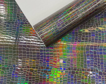 Op de afbeelding: Een rol iriserend imitatieleer met een krokodillenleerpatroon. Het materiaal reflecteert een regenboog aan kleuren, waaronder groen, blauw en paars. Het materiaal is gedeeltelijk opgerold en toont een witte achterkant.