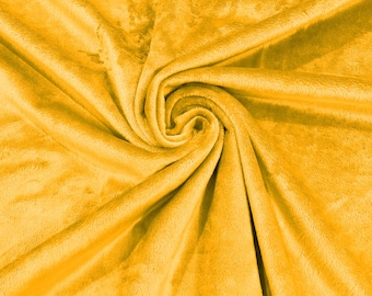 Puede incluir: Una manta de tela de felpa en color amarillo mostaza vibrante con una textura suave y aterciopelada. El material está recogido en un remolino central, creando pliegues profundos y sombras que resaltan la densidad del tejido.
