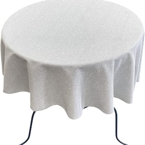 May include: A round, white tablecloth with a subtle sparkle finish. The tablecloth is draped over a round table with black metal legs.