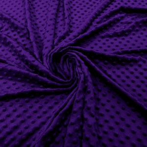 Puede incluir: Primer plano de una tela morada oscura con un patrón texturizado de puntos. La tela es suave y afelpada, con un ligero brillo. El material está recogido en espiral, creando sombras y resaltando la textura.
