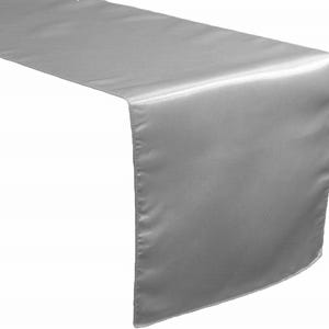 May include: A silver satin table runner. The runner is draped over a surface, showcasing its smooth, reflective texture. The fabric has a subtle sheen, adding a touch of elegance. Perfect for weddings, parties, or home decor.