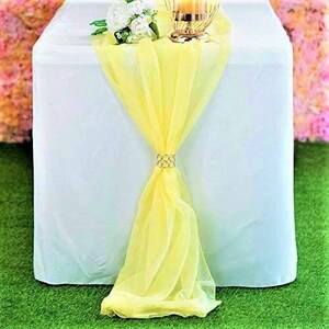 May include: A yellow sheer table runner with a gold ring accent on a white tablecloth. The runner is draped over the table and hangs down on both sides.