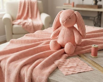 Puede incluir: Un peluche de conejo color melocotón está sentado sobre una manta a juego de textura suave. Un pequeño trozo de tela cuadrado, un carrete de hilo a juego y una regla de madera están colocados sobre la superficie de madera junto al peluche.