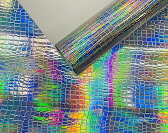 Op de afbeelding: Een rol iriserend, getextureerd vinyl met een krokodillenleerpatroon. Het materiaal reflecteert een spectrum aan kleuren, waaronder blauw, groen, geel en paars. Het oppervlak heeft een glanzend, holografisch effect.