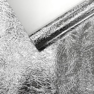 May include: A close-up of a silver metallic foil with a crinkled texture. The foil is partially rolled, revealing a smooth, white surface. The reflective material catches the light, creating highlights and shadows.