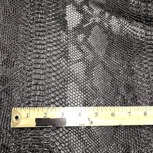 May include: Black faux leather fabric with a snakeskin pattern. A ruler is placed on the fabric to show the scale. The ruler measures 8 inches.