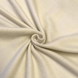 Puede incluir: Primer plano de una tela de color crema con una textura suave y peluda. La tela está doblada y drapeada, mostrando su tacto suave y lujoso.