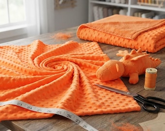 Puede incluir: Una tela minky de color naranja brillante con textura de puntos en relieve, una manta naranja doblada, un juguete de dragón de peluche naranja, un carrete de hilo naranja y una cinta métrica blanca con medidas en centímetros sobre una superficie de madera.