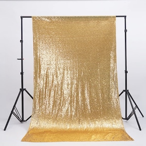 May include: Gold sequin backdrop for photography. The backdrop is hanging from a black stand and is made of shiny gold sequins.