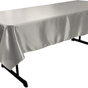 以下が含まれることがあります： 黒い折りたたみテーブルにかけられた、長方形のシルバーサテンのテーブルクロス。生地は滑らかで反射性があり、柔らかいハイライトと影を作り出しています。テーブルの長さは約180cmです。
