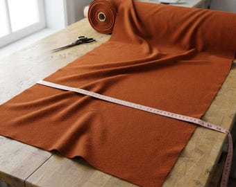 May include: A roll of burnt orange fabric with a textured surface, partially unrolled across a wooden table. A pair of black scissors rests on the fabric, and a white measuring tape is stretched across the width, showing a measurement of 58 inches.