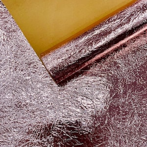 May include: A close-up of a roll of metallic, textured, rose-colored paper with a gold-colored sheet on top. The paper has a crinkled appearance, reflecting light. The image suggests a craft or decorative material.