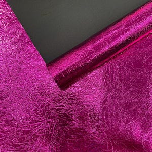 May include: A roll of vibrant hot pink material featuring a highly reflective, crinkled metallic texture. The material is partially unrolled, revealing a smooth, matte black underside that contrasts with the bright, textured surface.