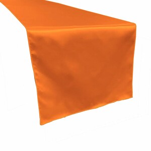 May include: An orange satin table runner. The runner is folded over and hangs down, showcasing its smooth, shiny texture. The color is a vibrant, solid orange, perfect for adding a pop of color to any table setting. Ideal for weddings, parties, or home decor.